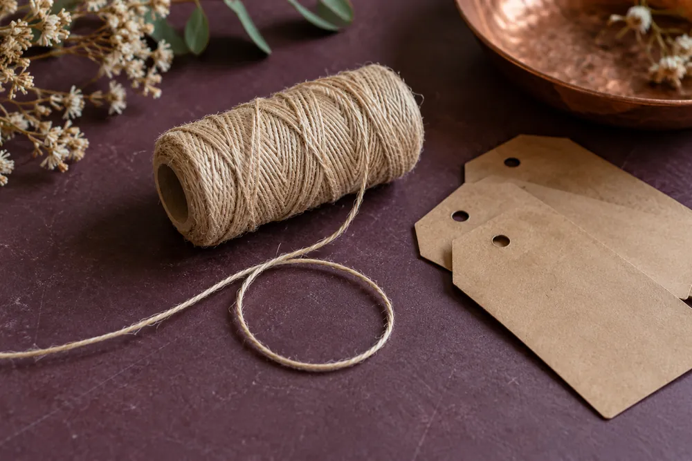 Ilustración del material: bobina de cordel fino natural junto a etiquetas kraft sin texto.